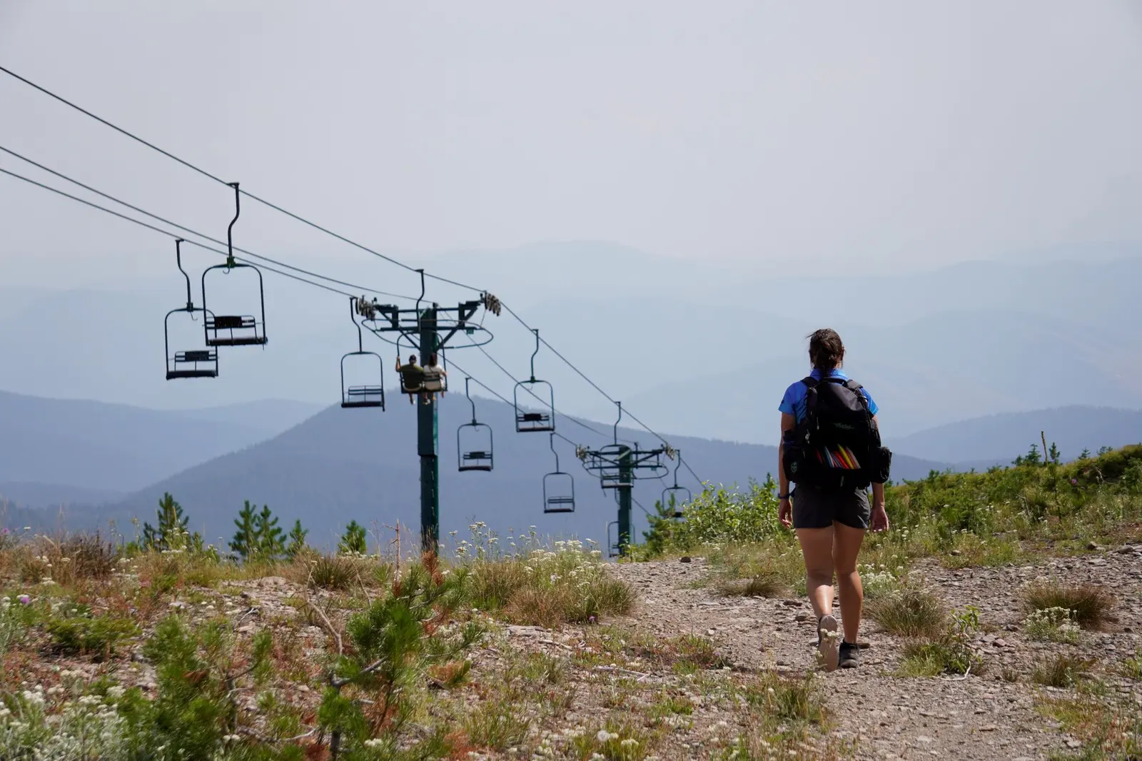 A person with a backpack walks on a mountain trail near a ski lift.
