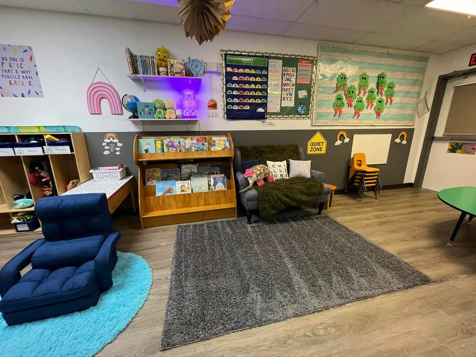 A colorful, well-organized early childhood classroom features a reading nook with a rug, chairs, and books on shelves.