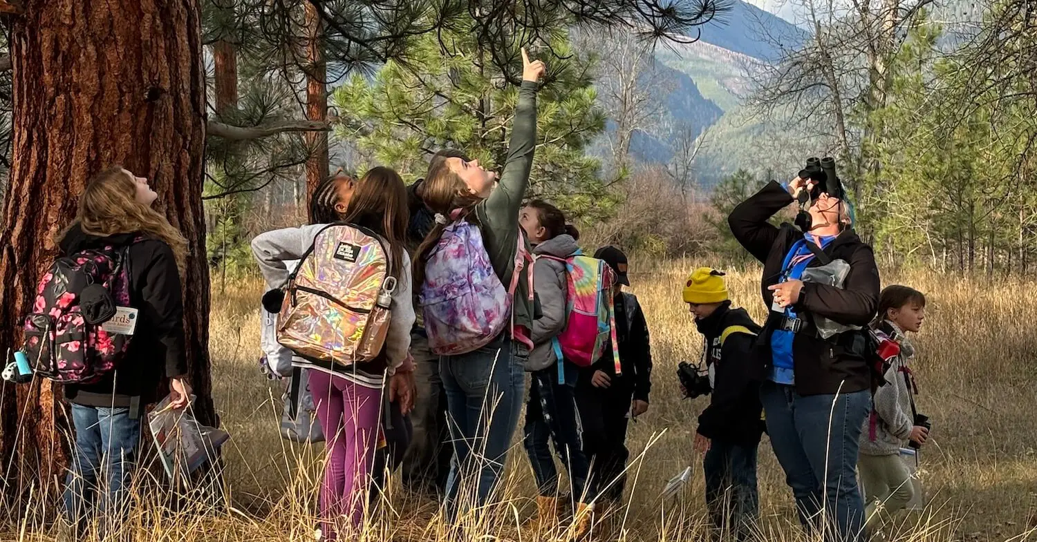 A group of children and an instructor participate in an outdoor birdwatching activity in a forest.