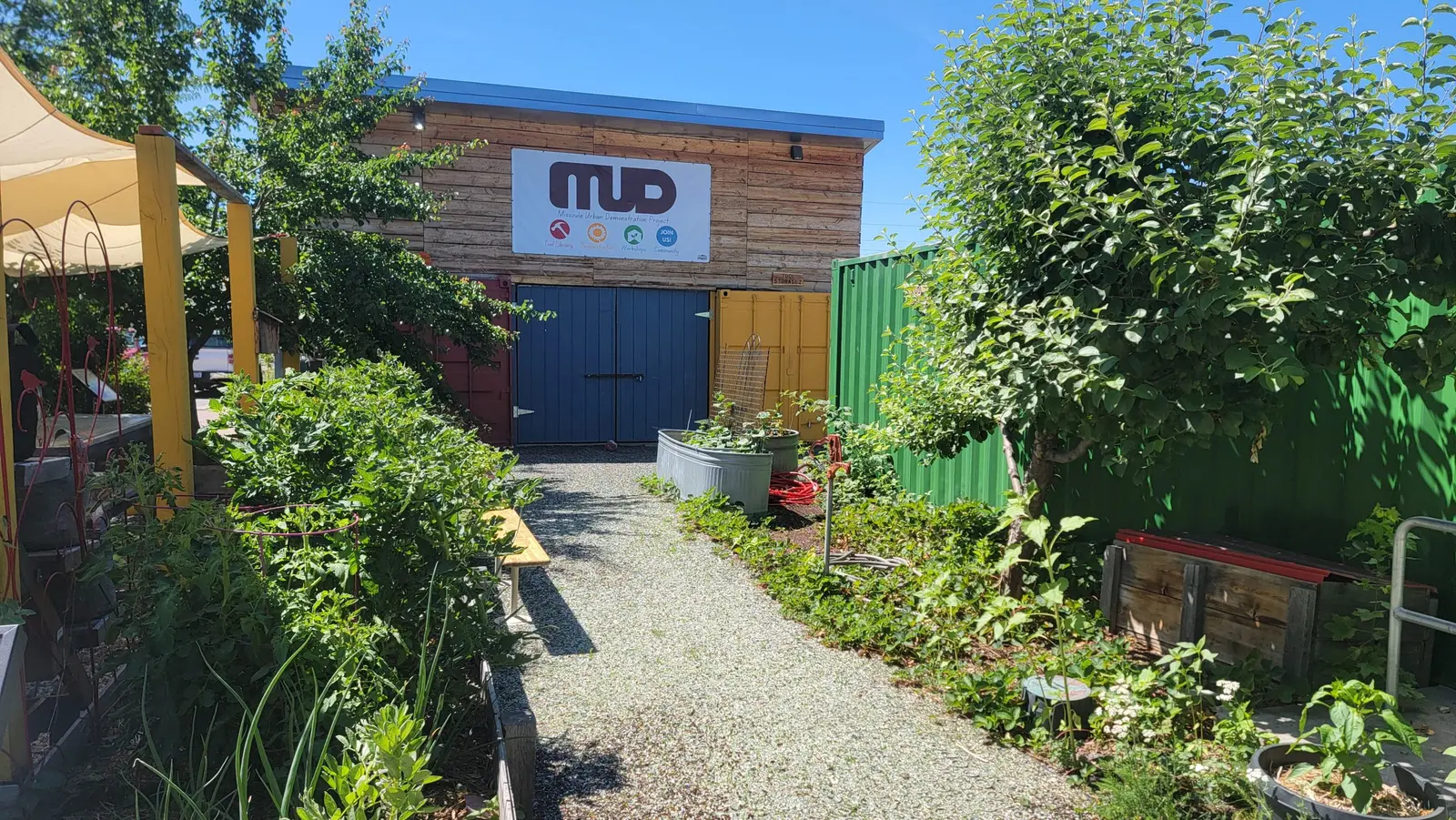 A gravel path leads to a wooden building with a MUD sign in a garden setting.
