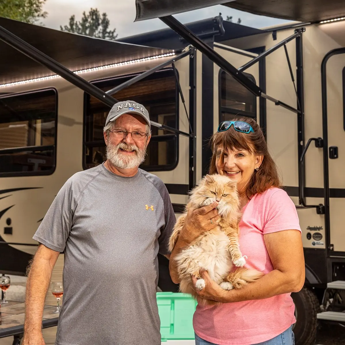An adult couple poses together while holding a fluffy ginger cat in front of a camper.