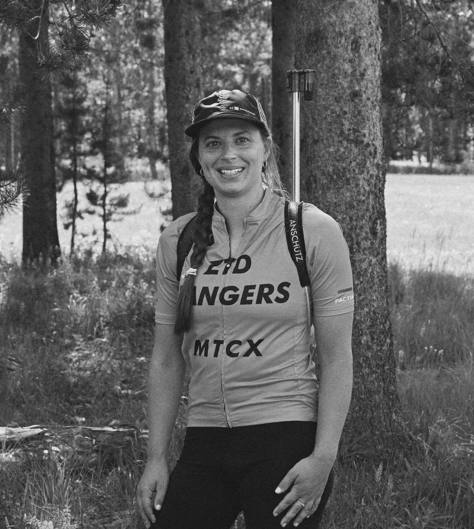 A woman stands in a forest wearing a jersey with text while carrying a rifle on her back.