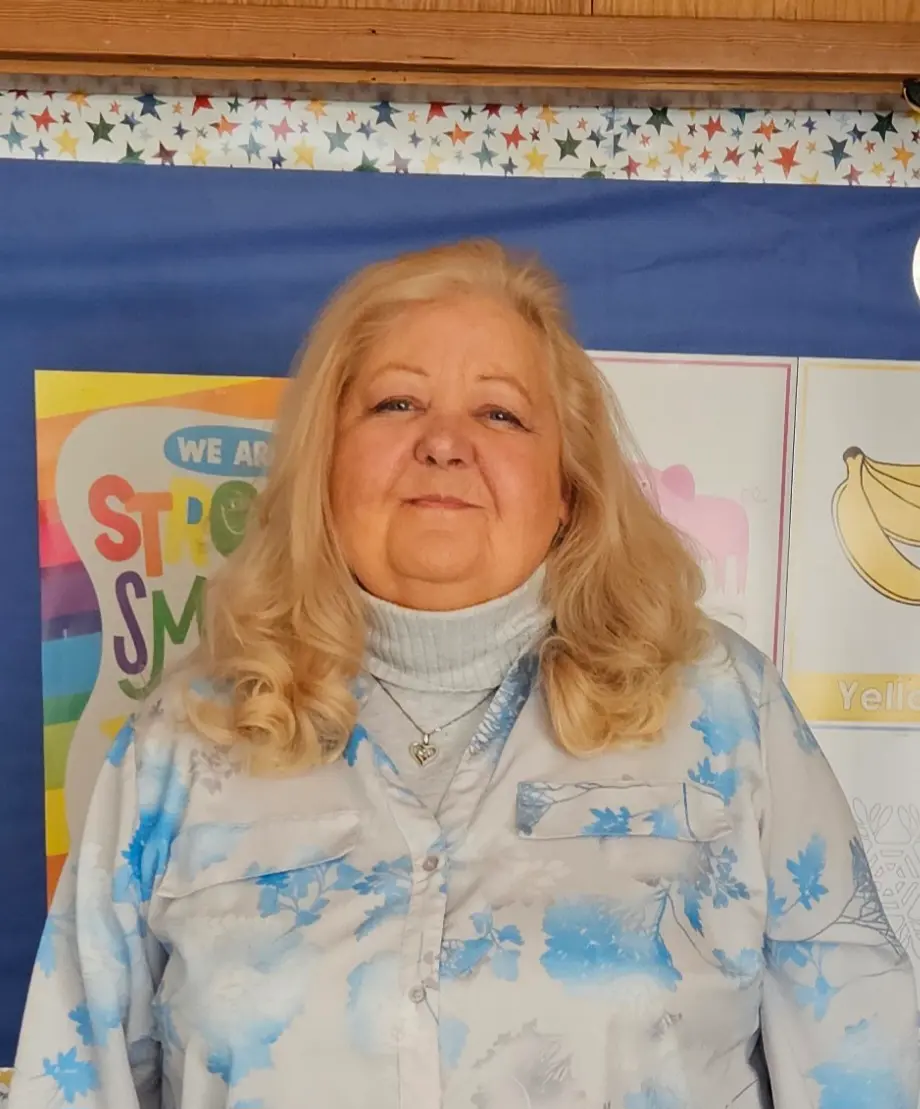 A woman with blonde hair stands in front of a bulletin board in what appears to be a classroom.