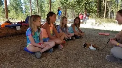 A group of children sits on the ground outdoors while observing a small animal on a leash held by an instructor.