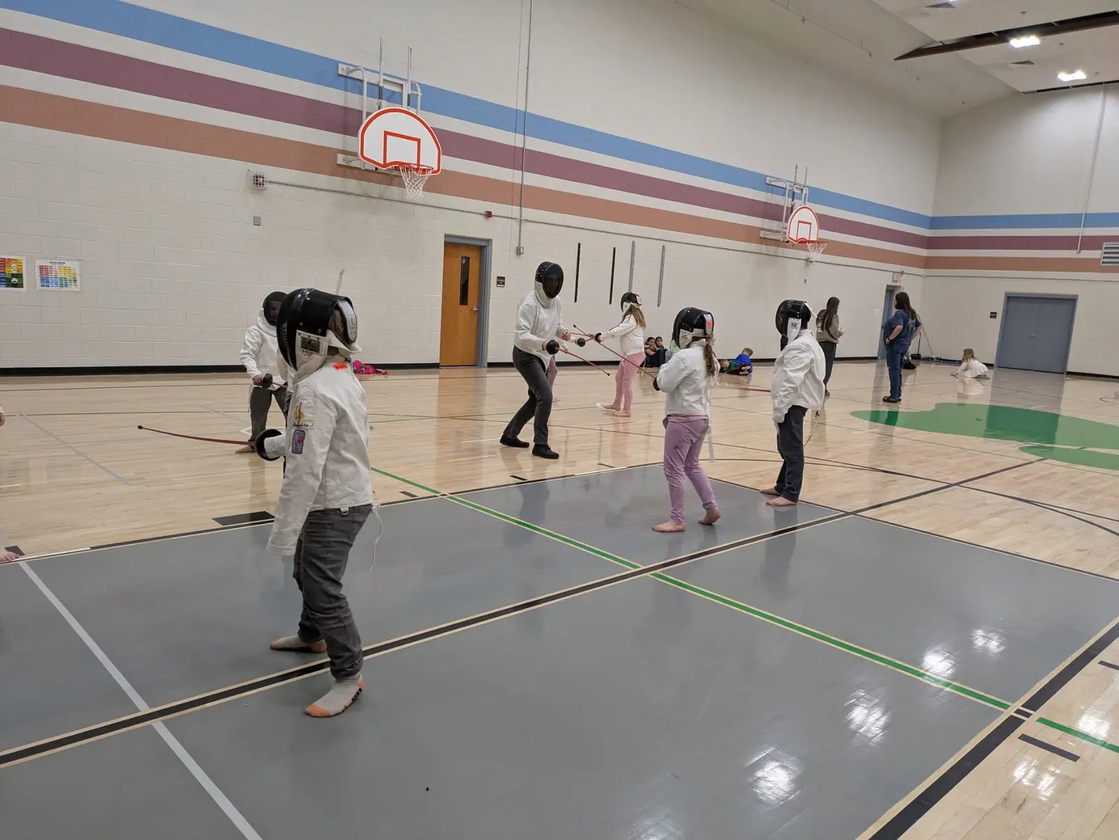 Children and an instructor are practicing fencing inside a gymnasium.