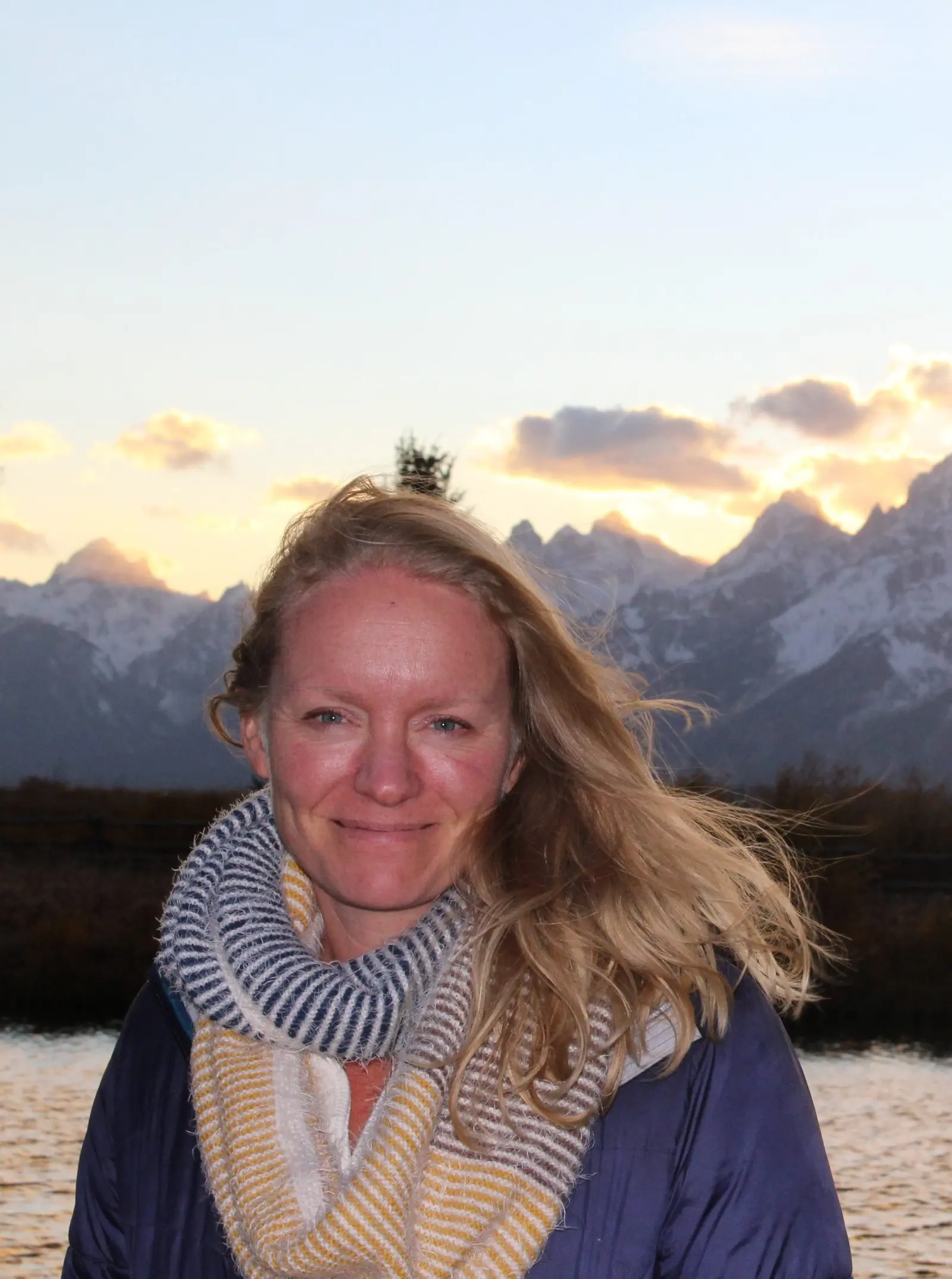A woman smiles in front of a mountain landscape at sunset.