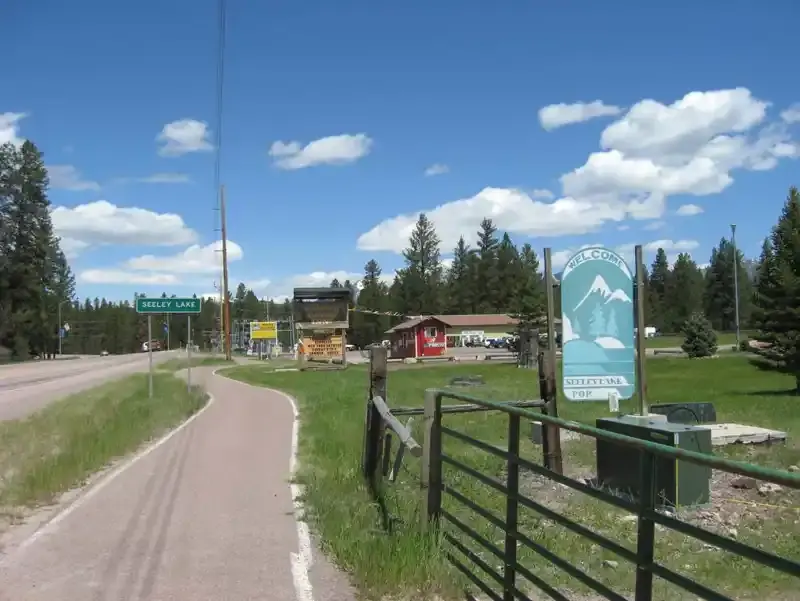 A paved path leads past a welcome sign and trees on a sunny day.