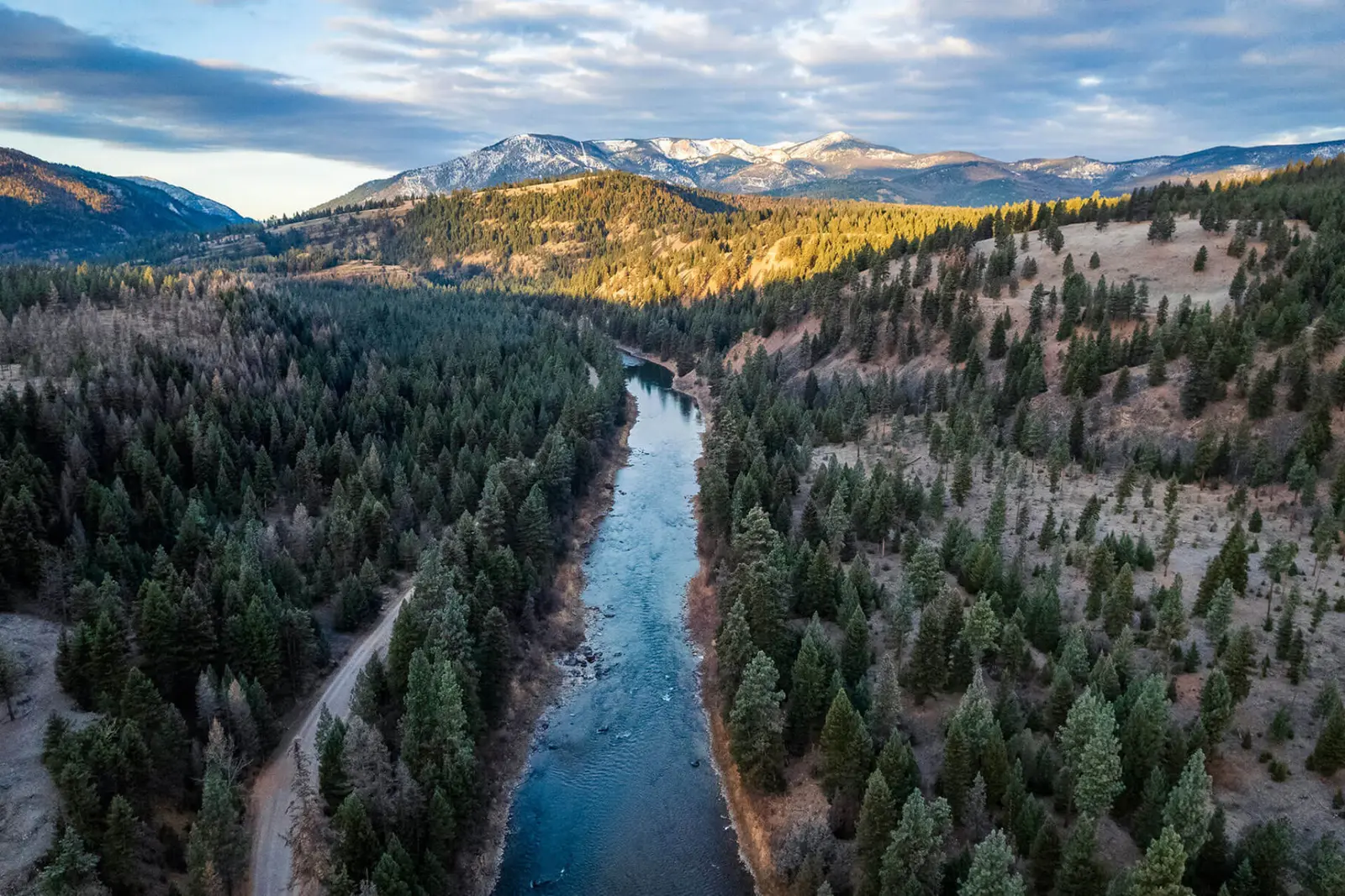 An aerial view of a winding river flowing through a forested valley with snow-capped mountains in the distance.