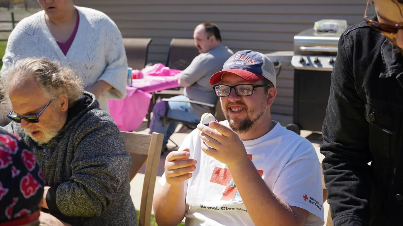 A man in a baseball cap smiles while holding a small egg during an outdoor gathering with adults.
