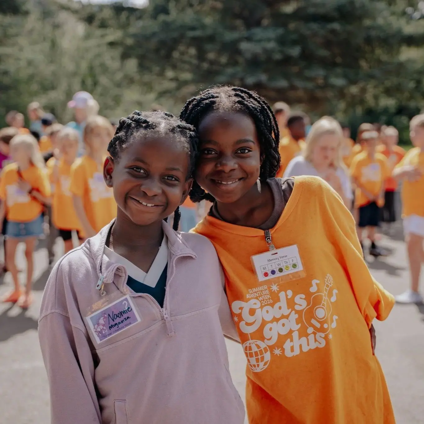 Two smiling girls stand together at an outdoor event where many other children in orange shirts are visible in the background.
