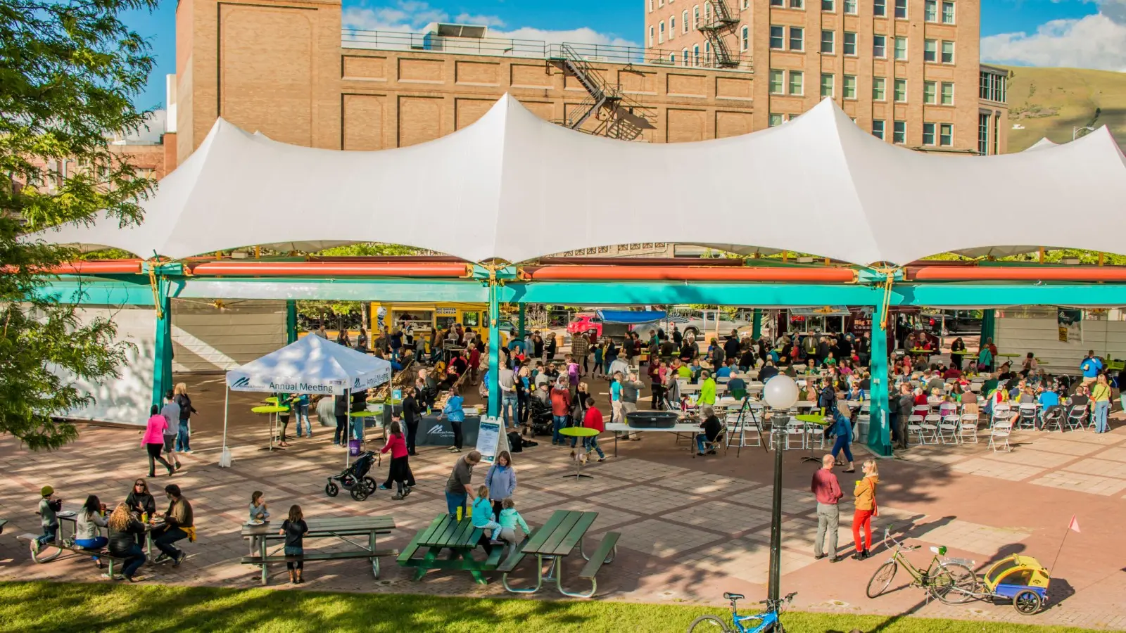 A large crowd of people gathers for an event in a public plaza under a large white canopy.