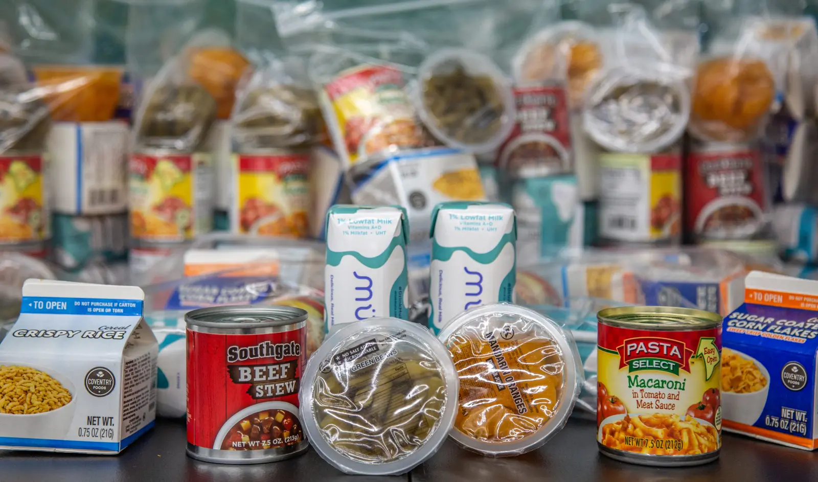 A collection of assorted non-perishable food items, such as canned goods, cartons, and plastic-sealed containers, are arranged together.