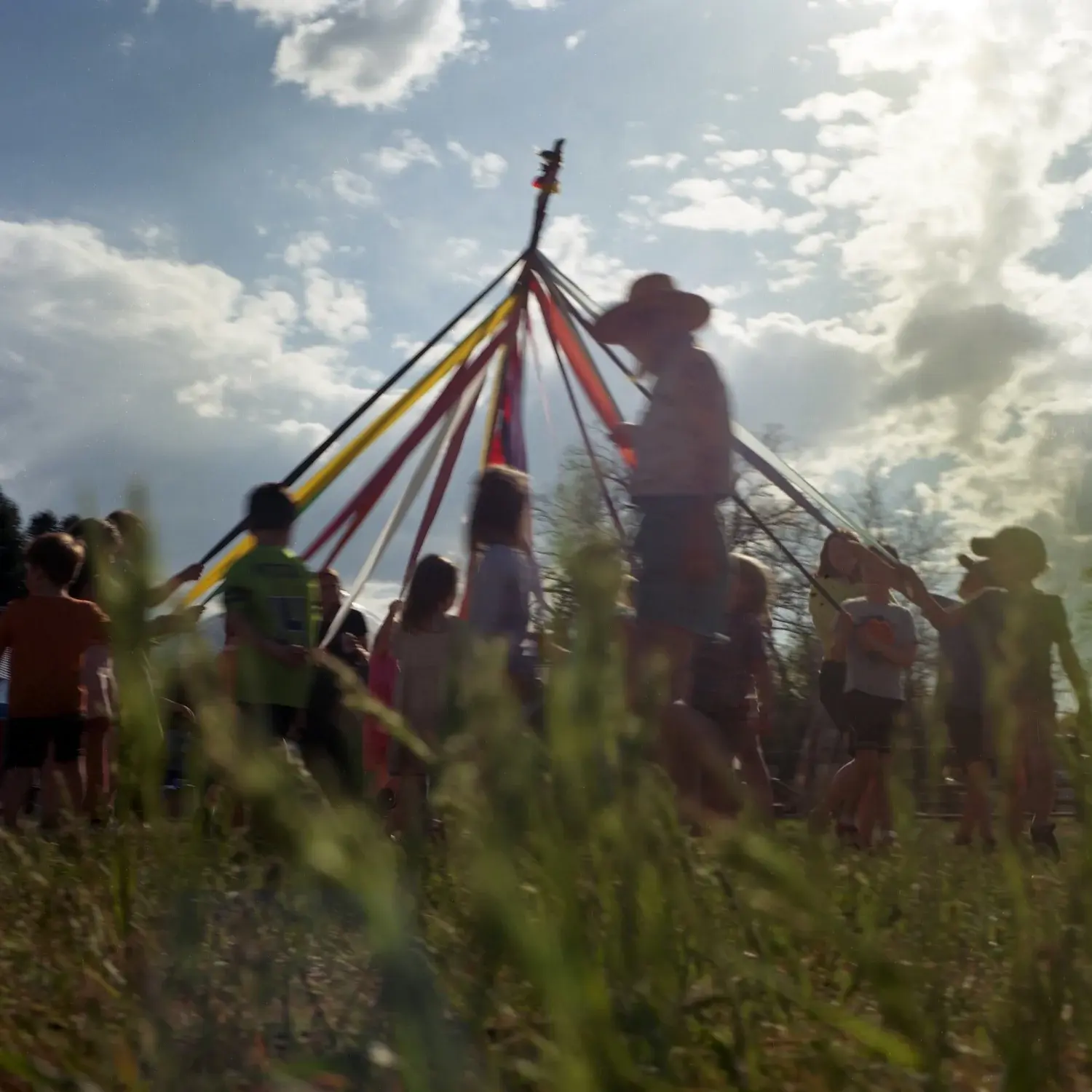 A group of children and an adult stand in a circle holding ribbons attached to a Maypole outdoors.
