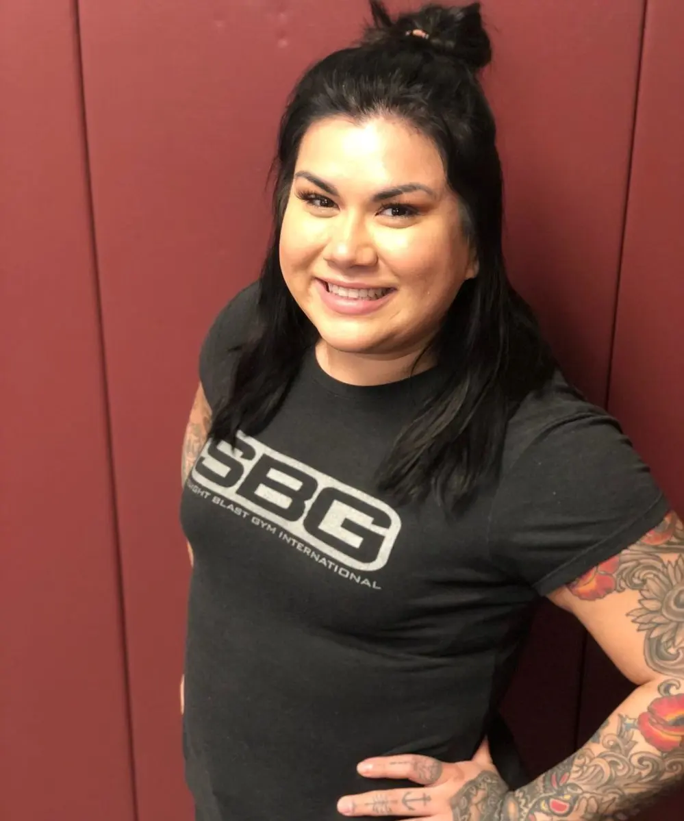 A smiling woman with tattoos stands against a maroon wall.