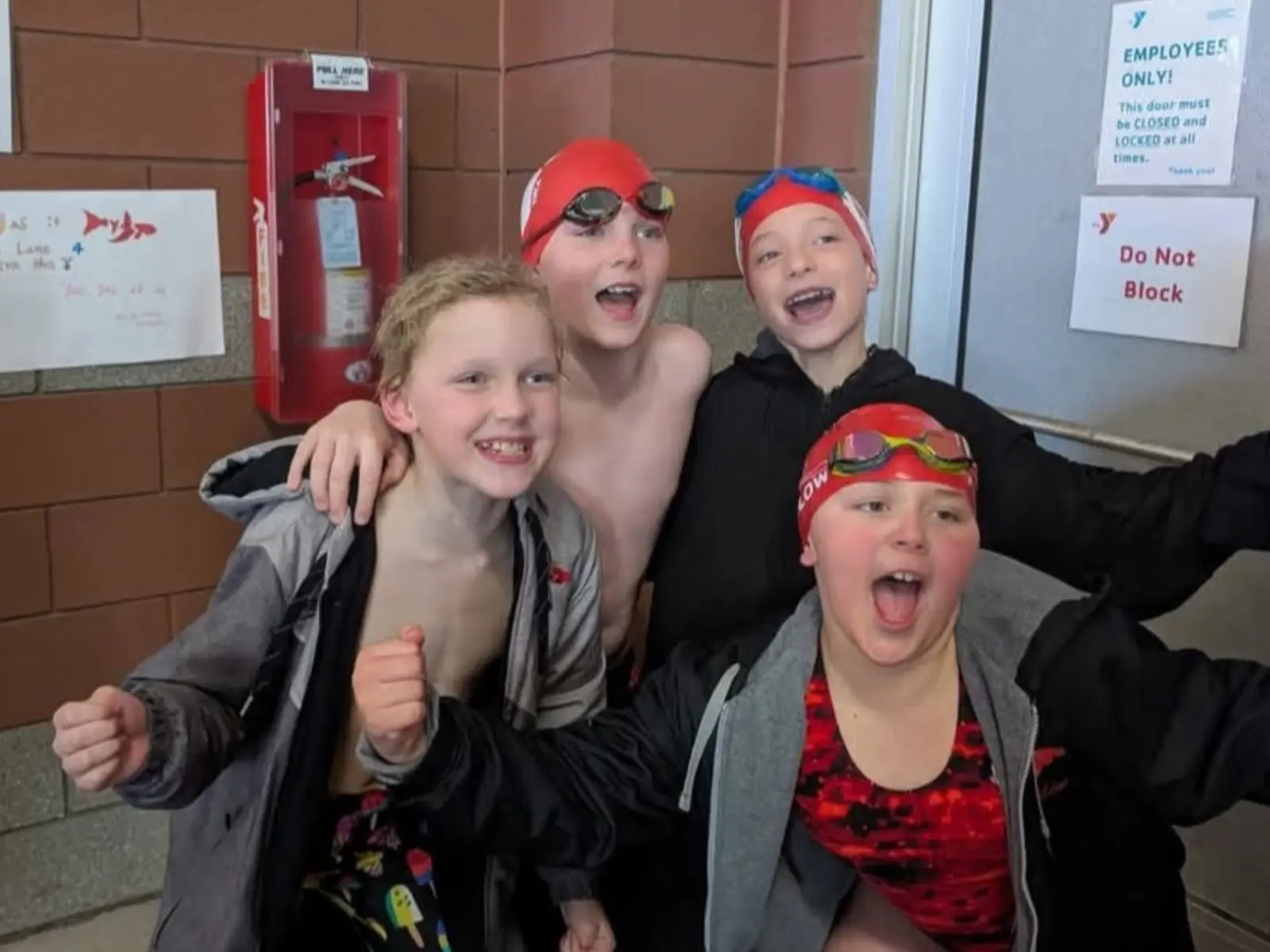 Four children in swim gear are smiling and cheering together indoors.