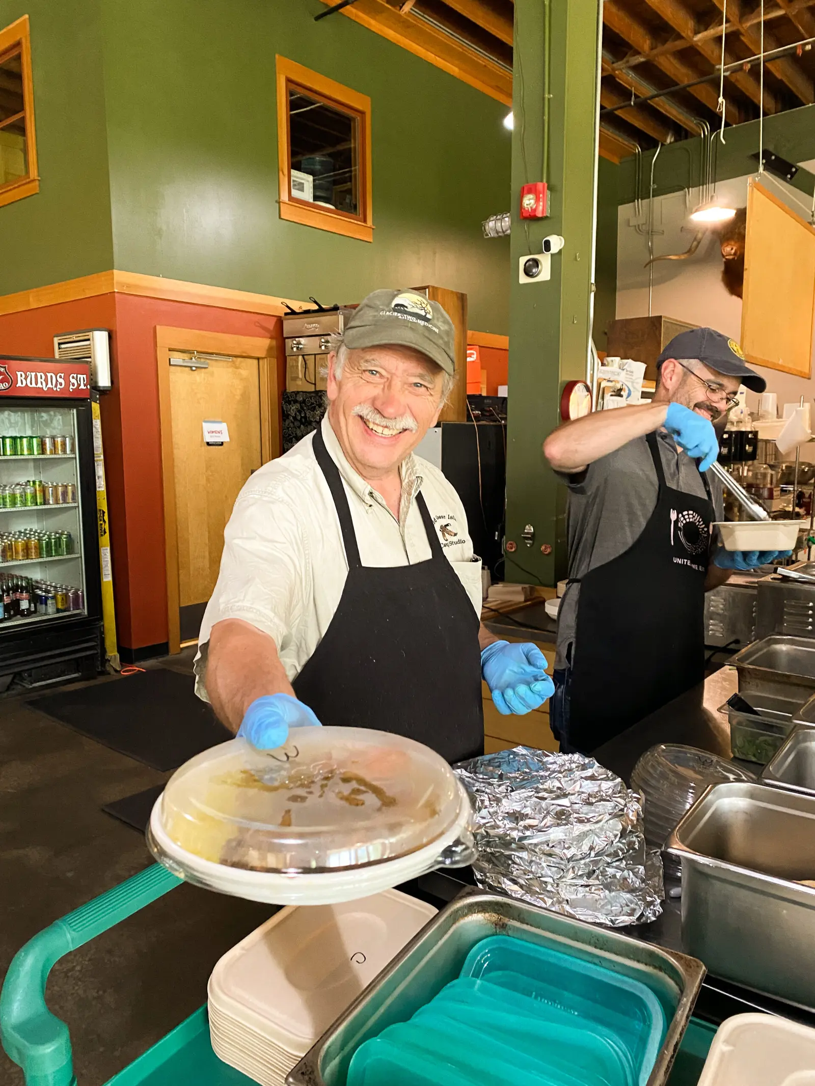 Two men wearing aprons and gloves prepare meals in a kitchen.