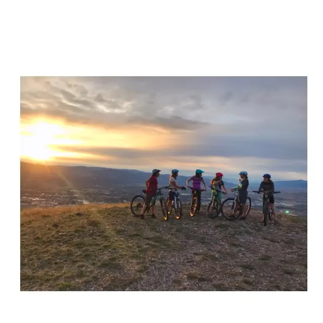 A group of cyclists stands with their mountain bikes on a hilltop overlooking a valley at sunset.