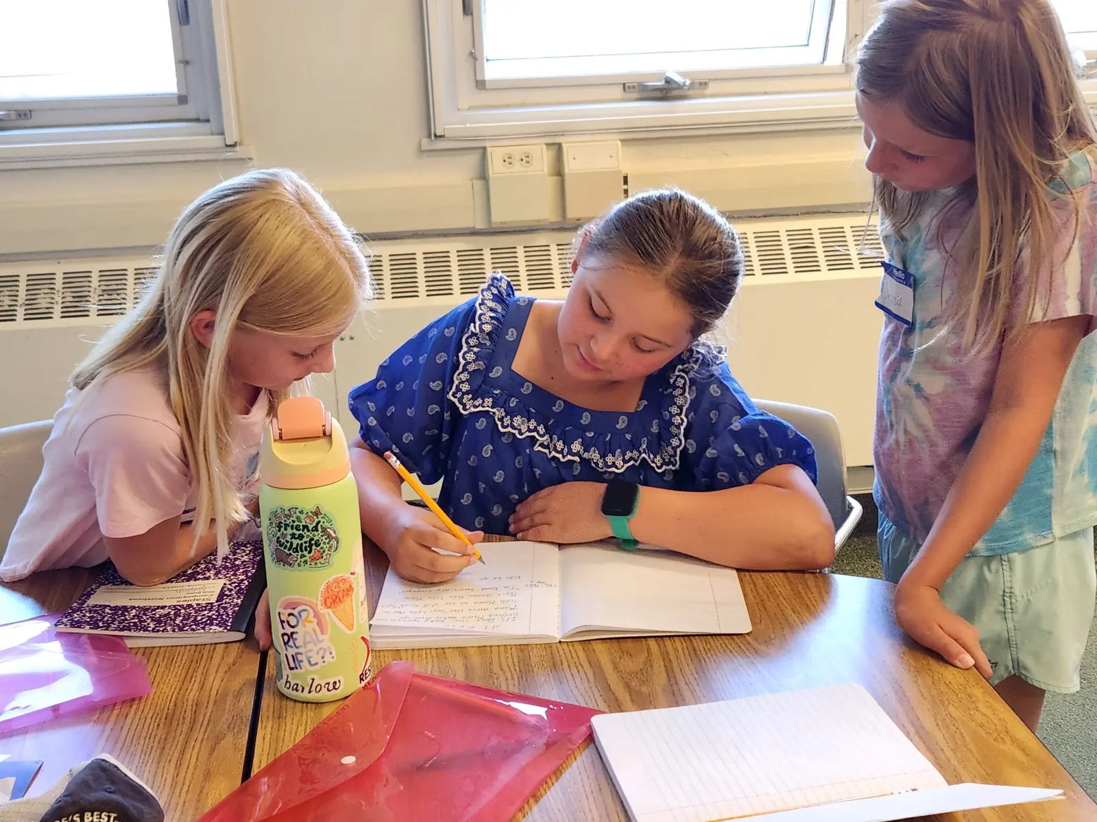 Three girls are working together on a writing activity at a desk in a classroom.