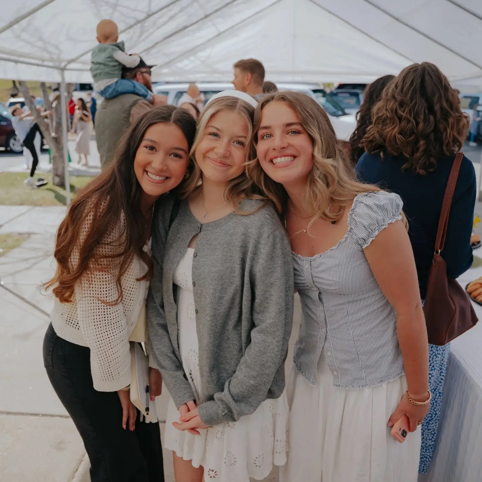 Three young women pose together for a photo under a white tent at an outdoor gathering.