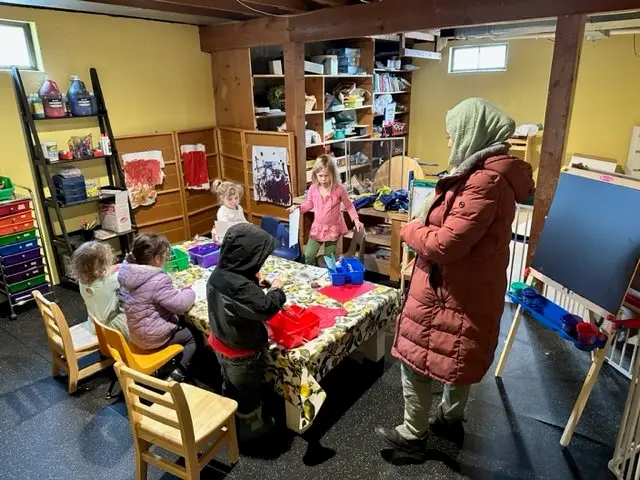 A teacher or caretaker with a hijab and winter coat supervising a group of children in a classroom. The children are sitting and standing around a table with art supplies, engaging in activities. The classroom has shelves, books, and art on the walls