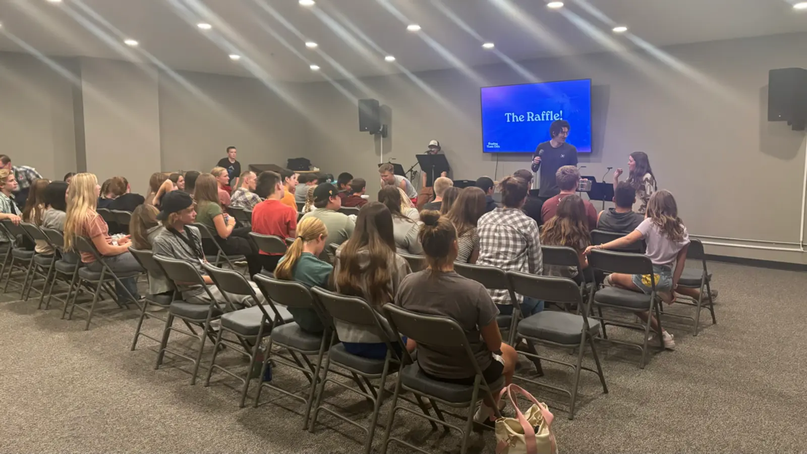 A large group of teenagers sits in chairs in a room while watching someone speak on stage during a raffle event.