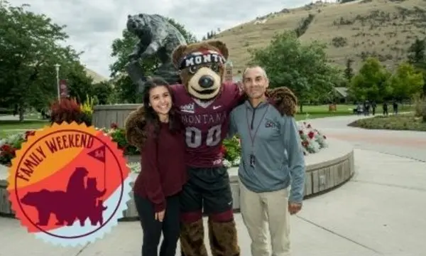 A young woman and an older man pose with a bear mascot in front of a campus landmark.