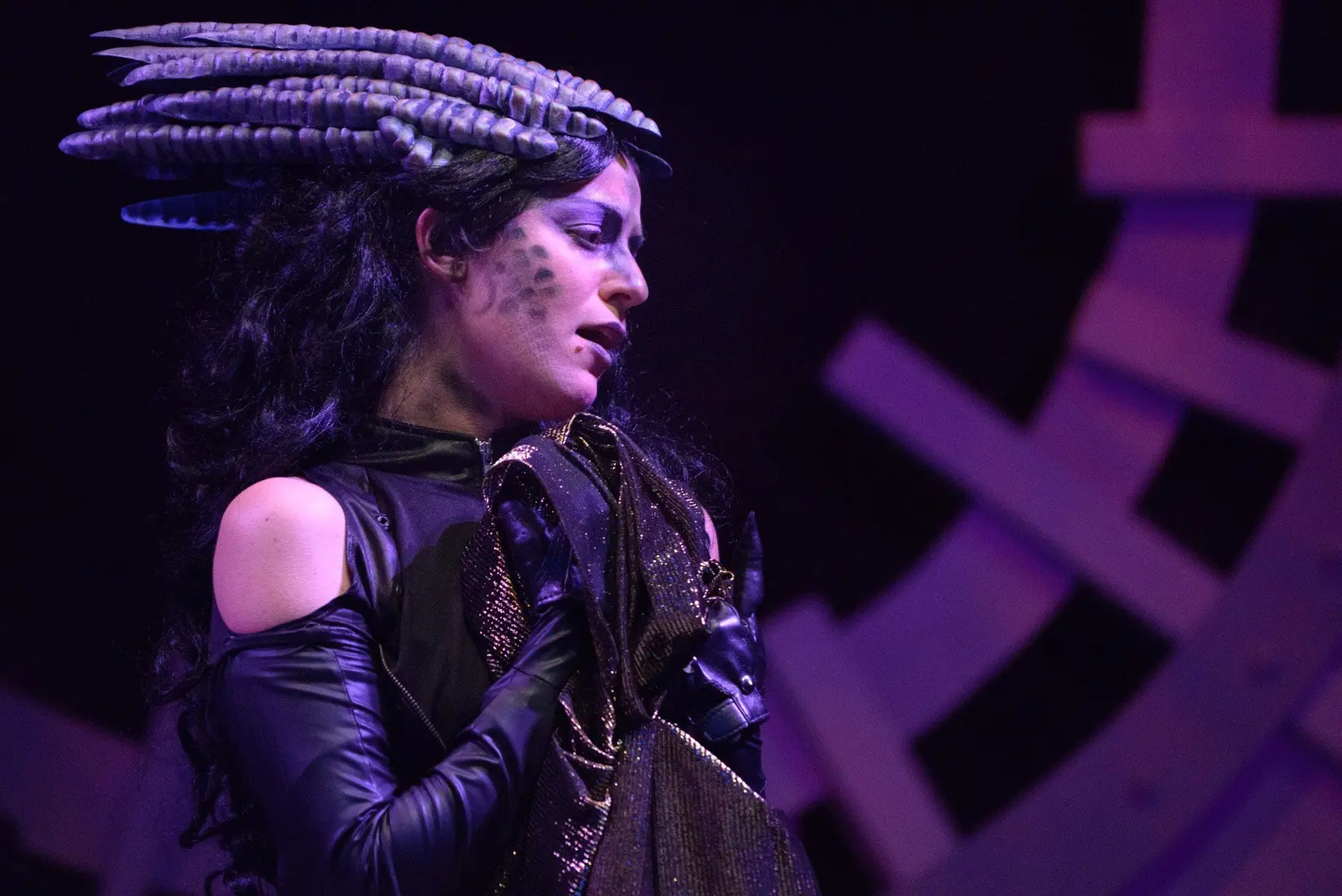 A woman dressed in a black costume with glitter and a headpiece with curled, striped horns. She has intricate face makeup resembling a mythical creature, with dark accents around her eyes and spots on her cheek. She is holding a shiny, metallic fabric close to her chest and appears to be in a theatrical performance, with a dark background and purple lighting.