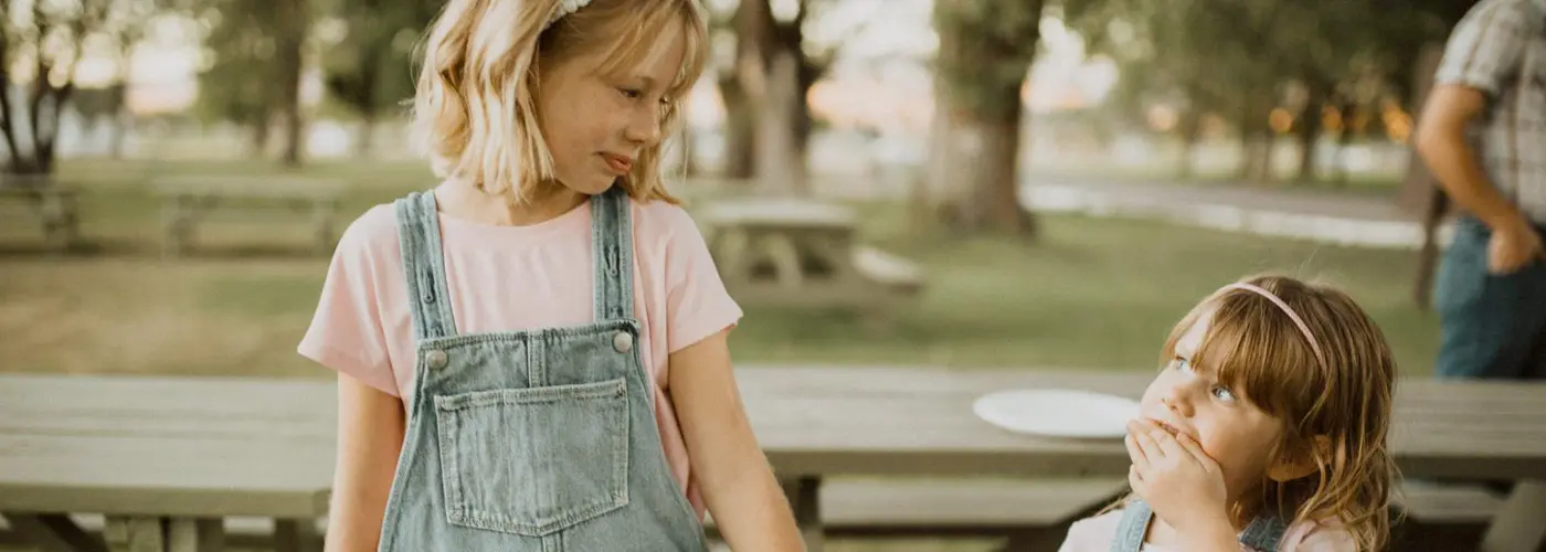 Two children in denim overalls interact at an outdoor picnic area.