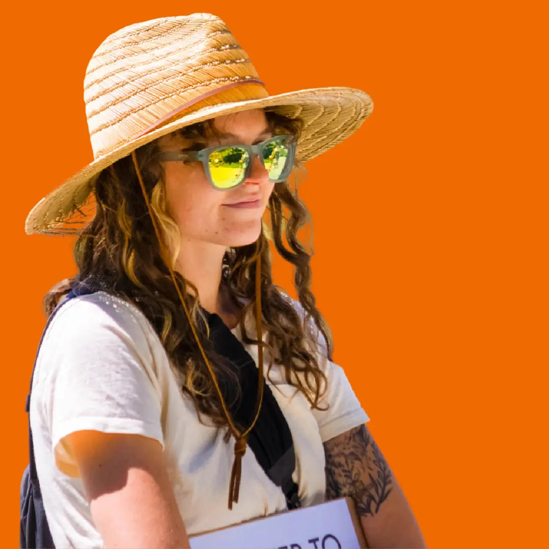 A woman wearing a straw hat and sunglasses poses against a bright orange background.