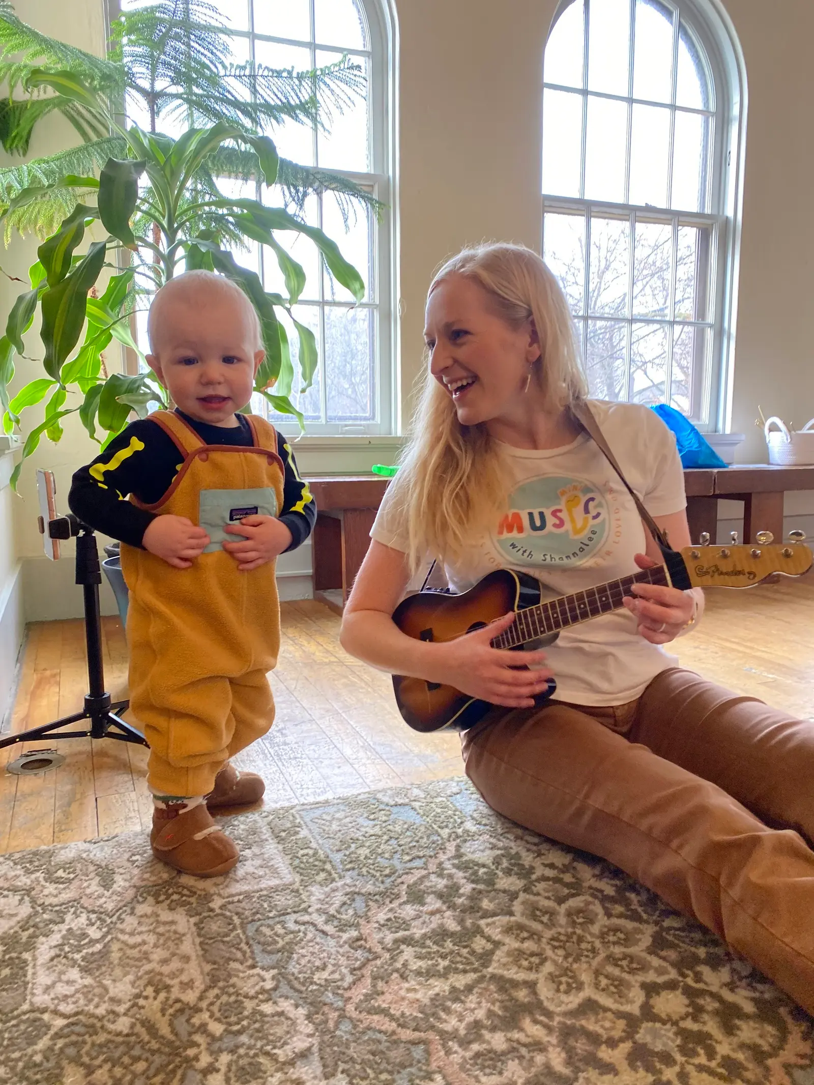 A woman plays a ukulele while a child stands nearby in a sunlit room.