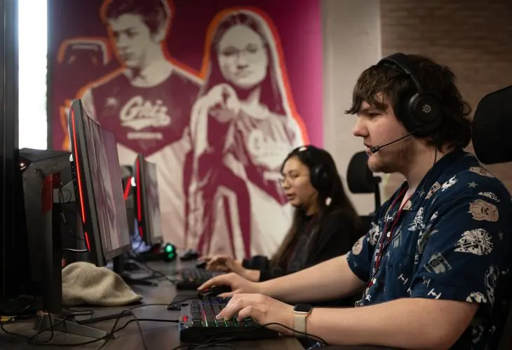 A young man sits at a gaming PC wearing a headset. His keyboard is lit with rainbow backlights. He looks intently at computer monitor.
