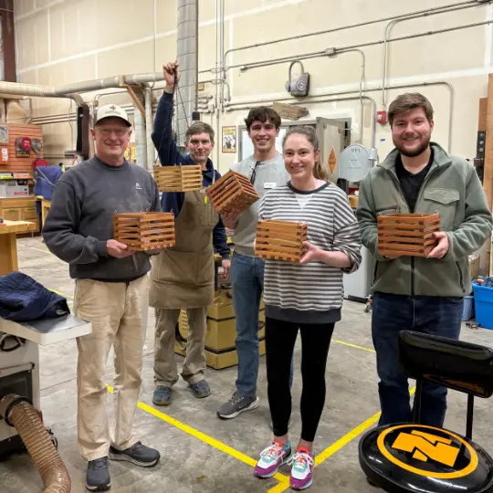 A group of adults in a workshop pose while holding wooden boxes they created.