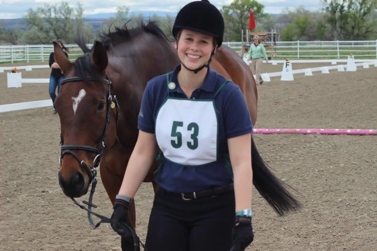 A smiling rider in equestrian gear stands beside her horse in an arena.