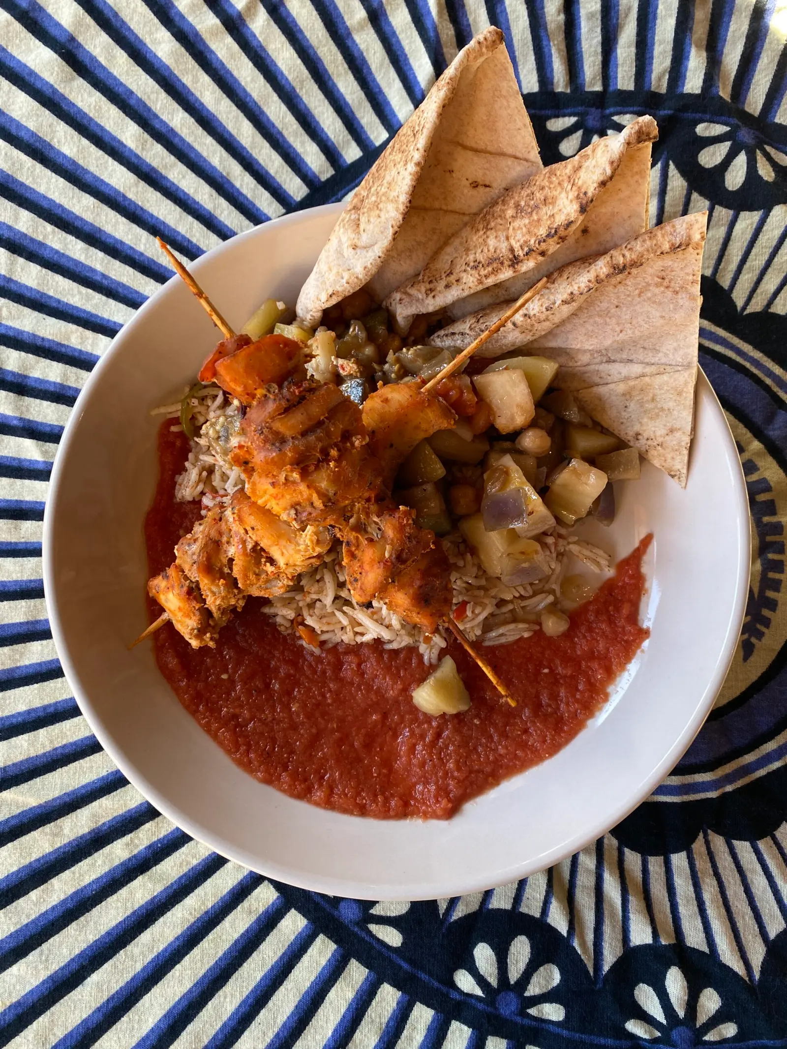 A bowl of skewered chicken with rice, vegetables, and pita bread is served on a patterned tablecloth.