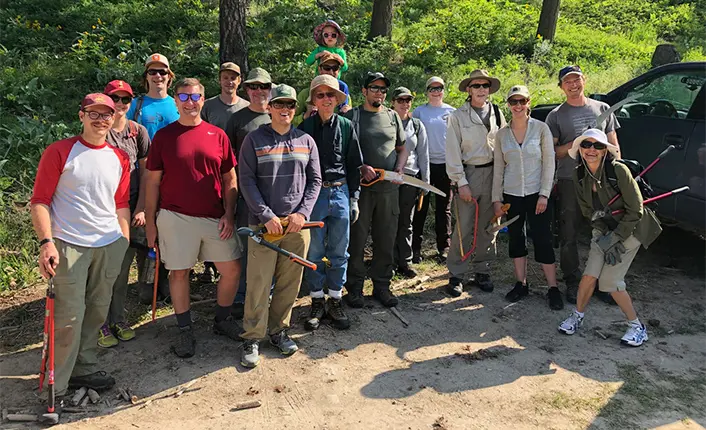 A group of volunteers poses for a photo with tools while working on a trail.