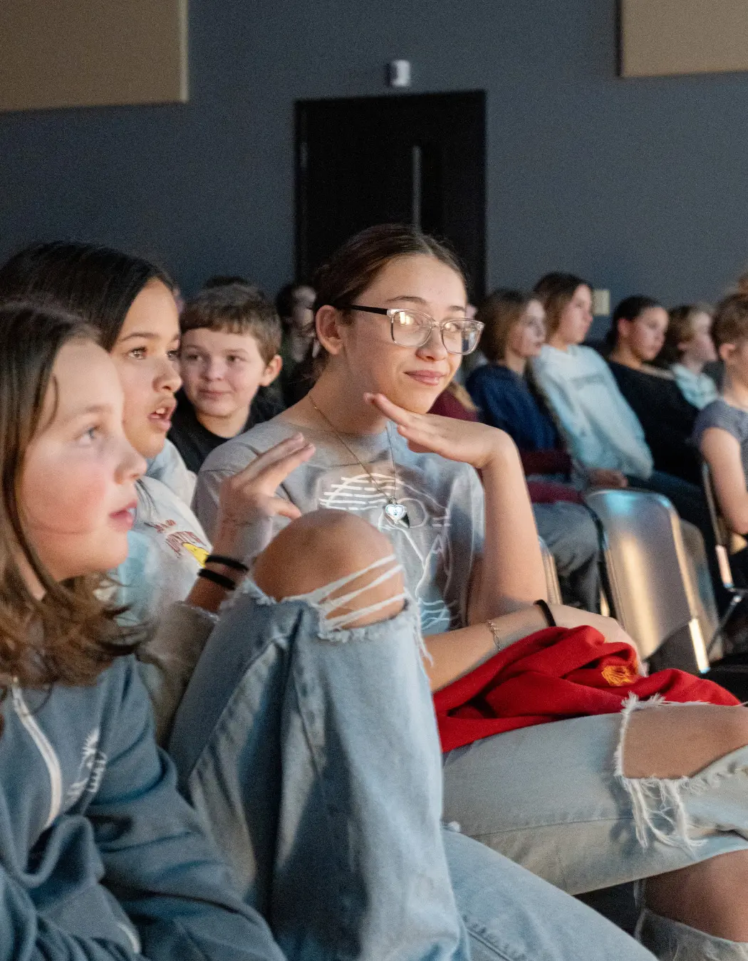 A group of children and teenagers sit in chairs, looking toward something off-screen.