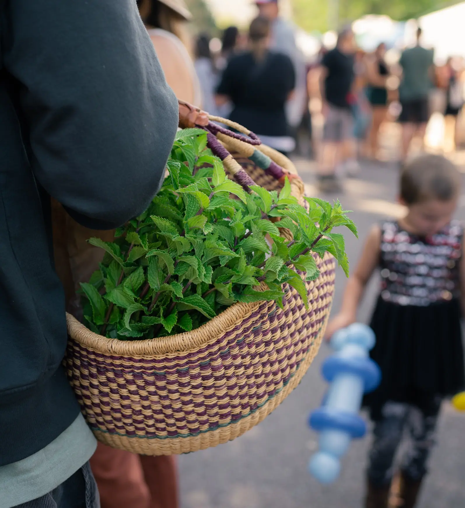 A person carries a basket filled with fresh mint through a crowded outdoor event.