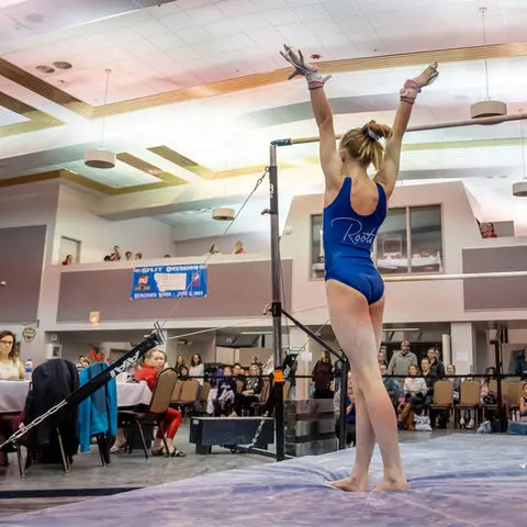 A gymnast in a blue leotard stands on a mat with her arms raised after a performance.
