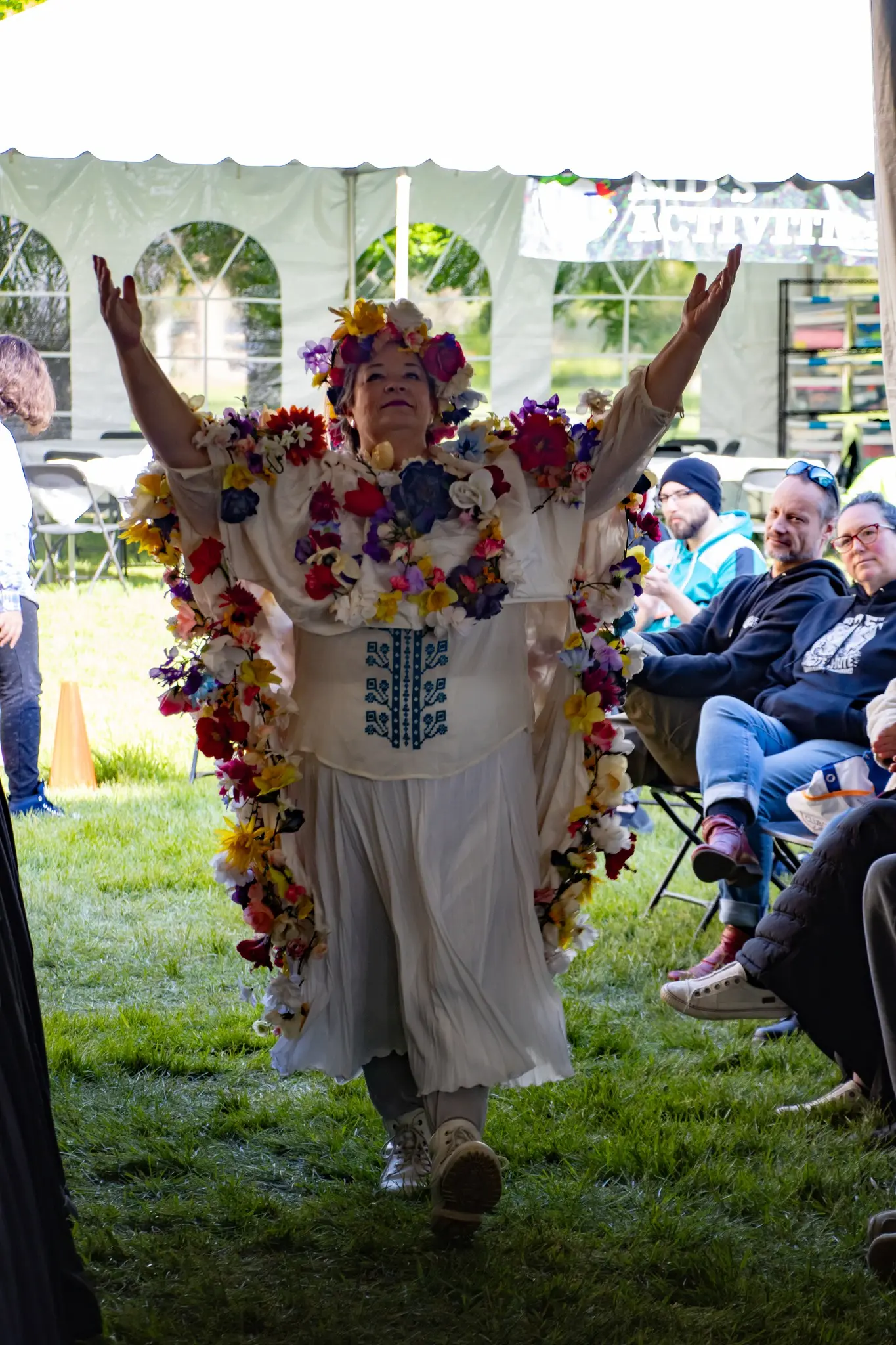 A woman wearing a flower-covered outfit performs for an audience seated under a tent.