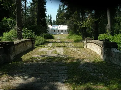 Photo by Forest Service Northern Region, CC BY 2.0 A weathered stone bridge leads toward a white building surrounded by trees.