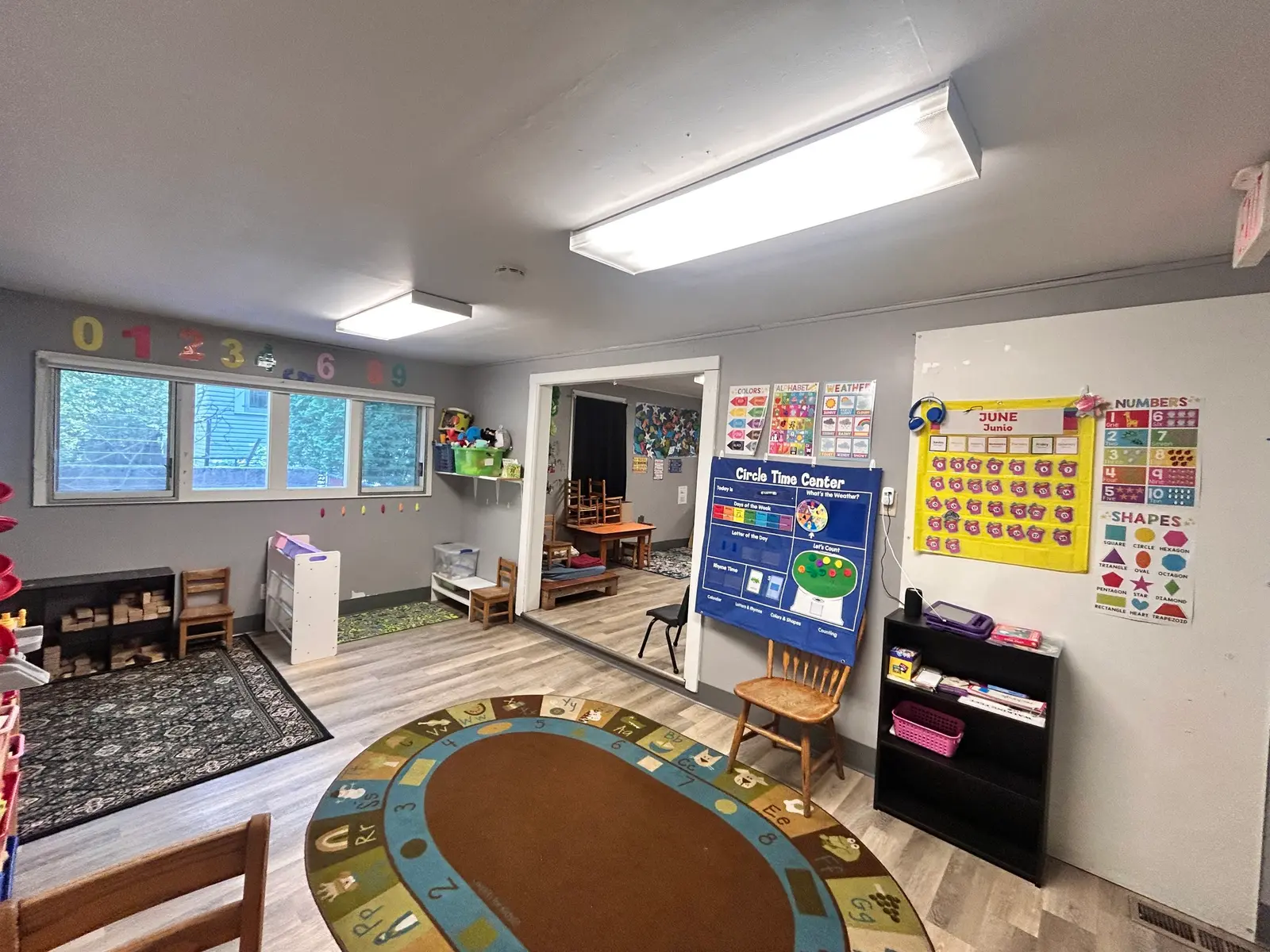 A bright classroom space is organized with educational materials, charts, and a circle-time rug.