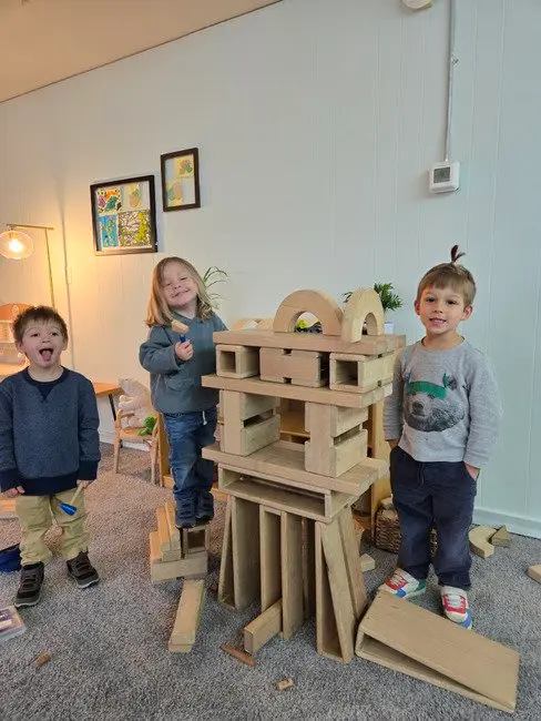Three young children pose while playing with large wooden building blocks in a room.