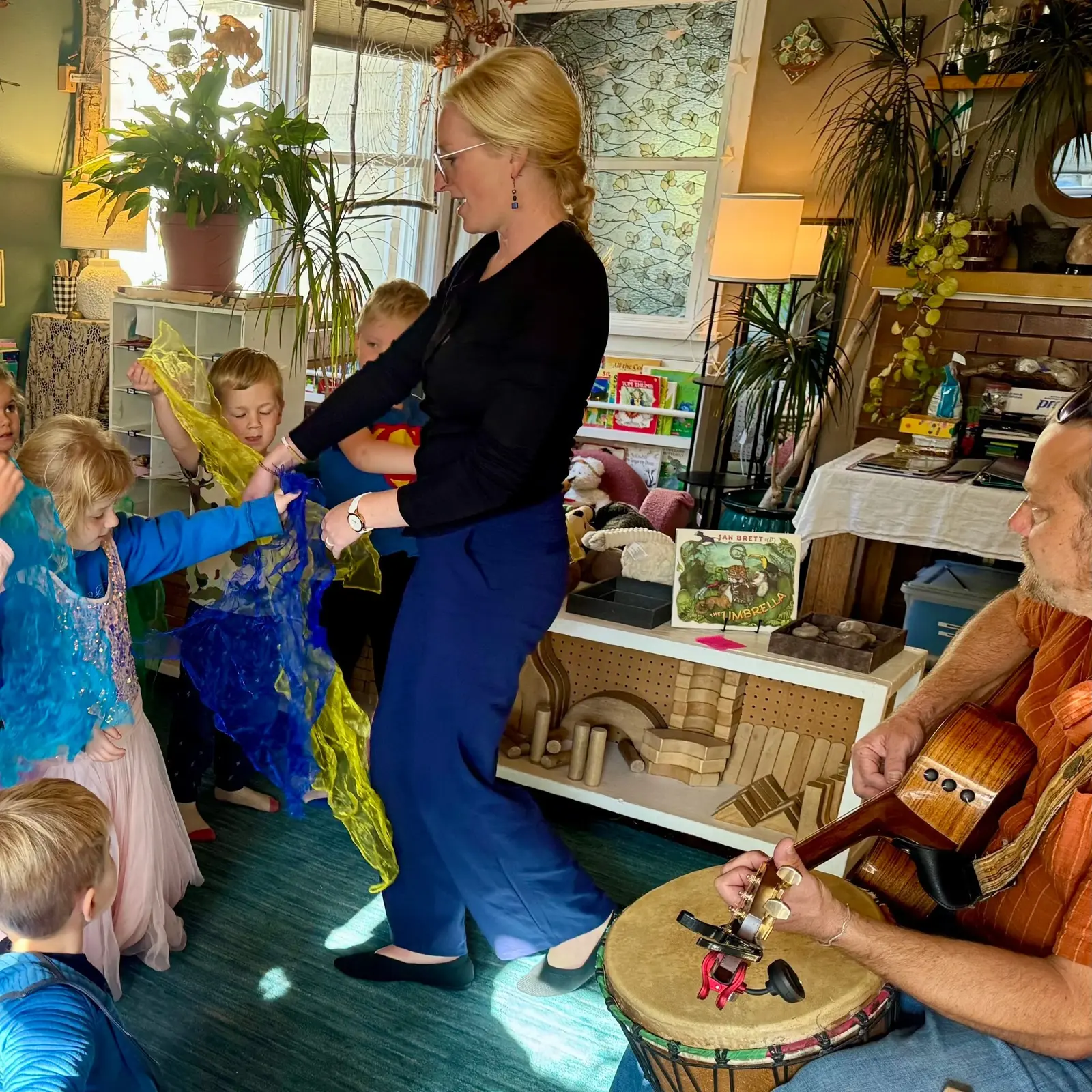 A woman leads a group of children playing with colorful scarves while a man accompanies them with music.