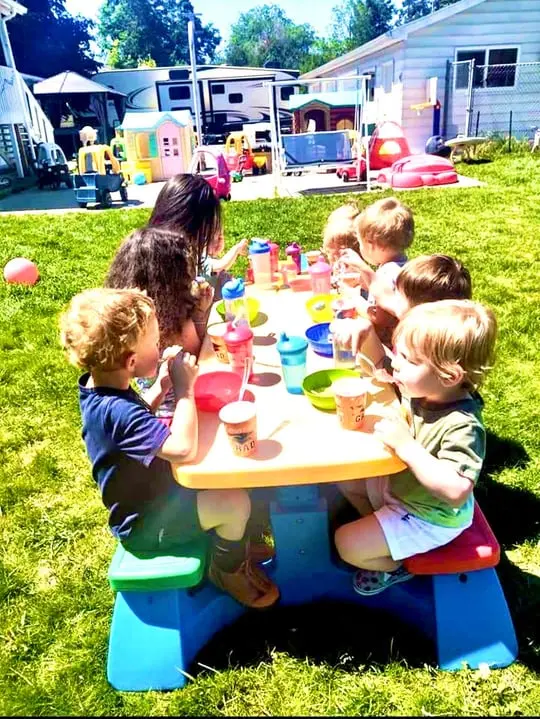 A group of young children sits at a plastic picnic table outdoors, enjoying snacks and drinks.