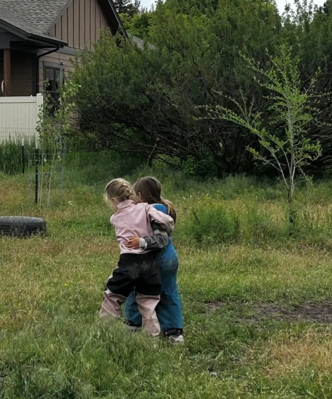 Two children in outdoor clothes hug each other while standing in a grassy field.