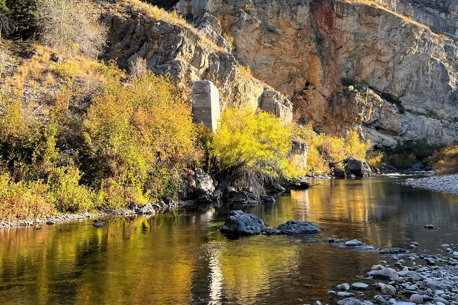 A quiet river flows alongside rocky cliffs and vibrant autumn vegetation with a concrete pillar standing near the water.