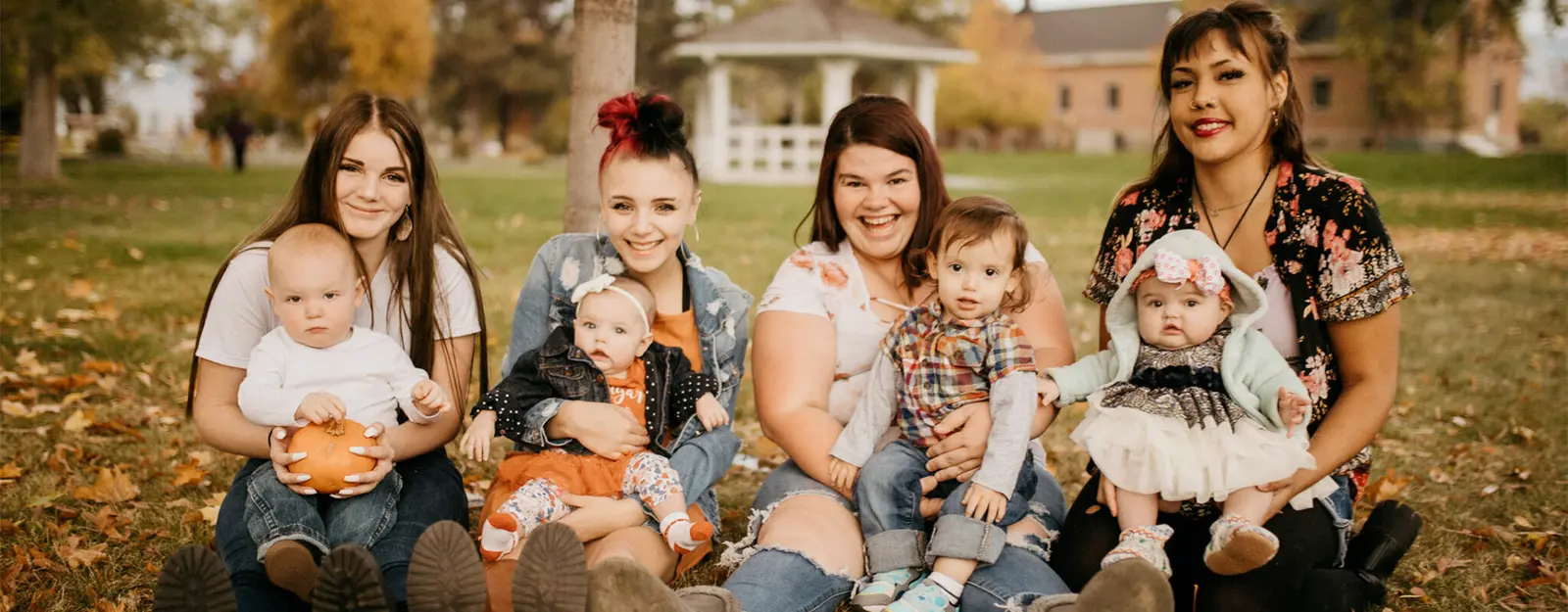 Four young women are sitting on a grassy field with their babies.