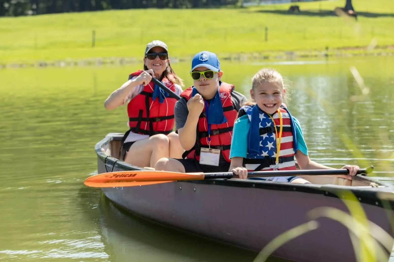 A woman and two children wearing life jackets paddle in a canoe on a pond.