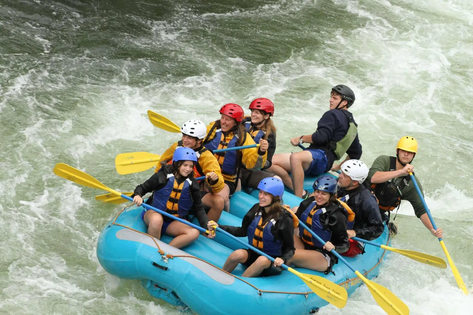 A group of people wearing helmets and life vests paddles a raft through white-water rapids.