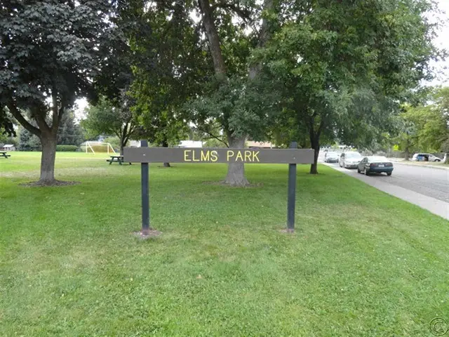 A park sign stands in a grassy area with trees and a road nearby.